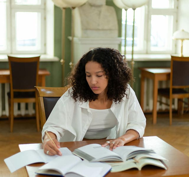 A woman with curly hair studies at a table, writing in a notebook with several open books in front of her, thoughtfully exploring our causes in a well-lit library or study room.