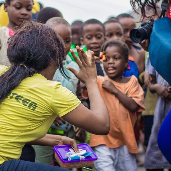 A woman in a yellow shirt faces a group of smiling children outdoors, holding a small container. One child reaches out towards her hand, while others watch and a photographer captures the moment—a heartwarming scene that beautifully reflects the impact of our causes.