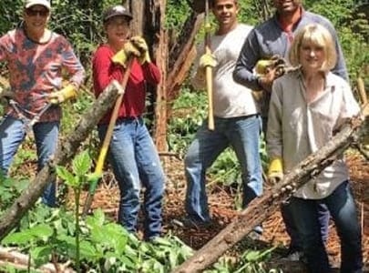 Five people wearing gloves hold tools and stand among cut branches in a wooded area, working together on an outdoor project organized by the Bellevue Breakfast Rotary Club.