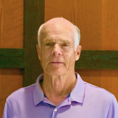 An older man with short, thin white hair wearing a light purple polo shirt stands in front of a wooden wall with a dark cross shape behind him.
