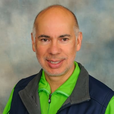 A man with a bald head and light facial hair smiles at the camera. He is wearing a green zip-up shirt under a dark blue vest. The background is a soft, blurred mix of blue and gray tones.