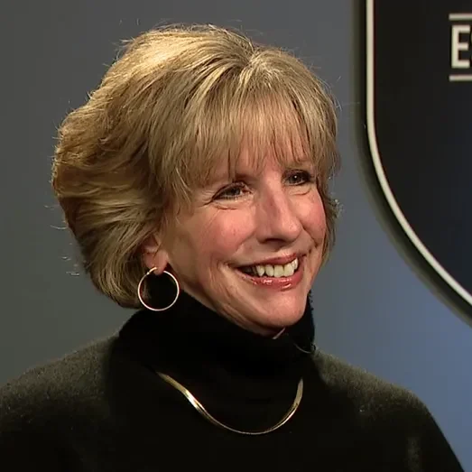 A smiling woman with short, light brown hair wearing hoop earrings, a black turtleneck, and a gold necklace, standing in front of a gray background with part of a black and white sign visible.