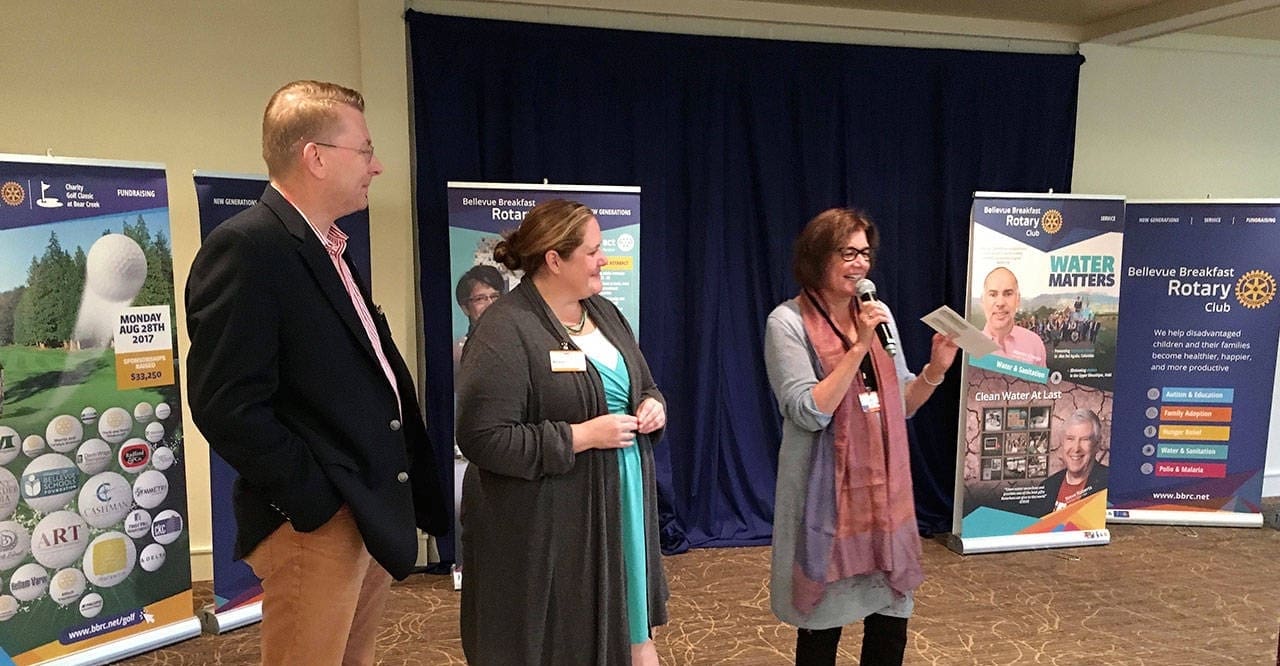 Three people stand and speak in front of Rotary Club banners at an indoor event. One woman holds a microphone and smiles, while the others listen, appearing engaged in conversation about upcoming events.