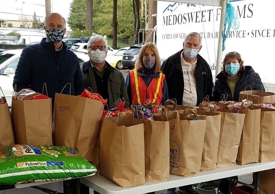 Five people wearing masks stand behind a table filled with grocery bags and pet supplies, likely distributing food or aid as part of a Bellevue Breakfast Rotary Club initiative. A trailer behind them reads MEDOSWEET FARMS. Trees and parked cars are visible in the background.