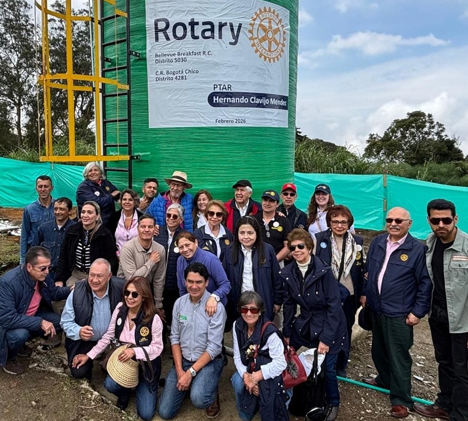 A group of people pose in front of a large green water tank with the Rotary logo and text, outdoors with trees and a green tarp in the background. Smiling and wearing casual or semi-casual attire, they proudly stand together to support our causes.