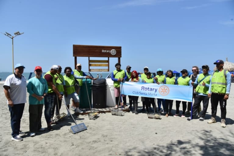 A group of people in green vests pose on a beach holding a Rotary Club Santa Marta banner, alongside members of the Bellevue Breakfast Rotary Club, with cleaning tools and trash bags, under a clear blue sky.