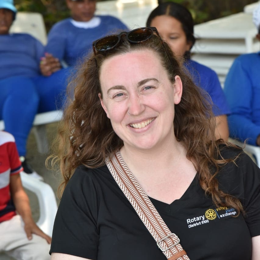 A smiling woman with light skin and long brown hair, wearing sunglasses on her head and a black Bellevue Breakfast Rotary Club shirt, sits outdoors with several people blurred in the background.