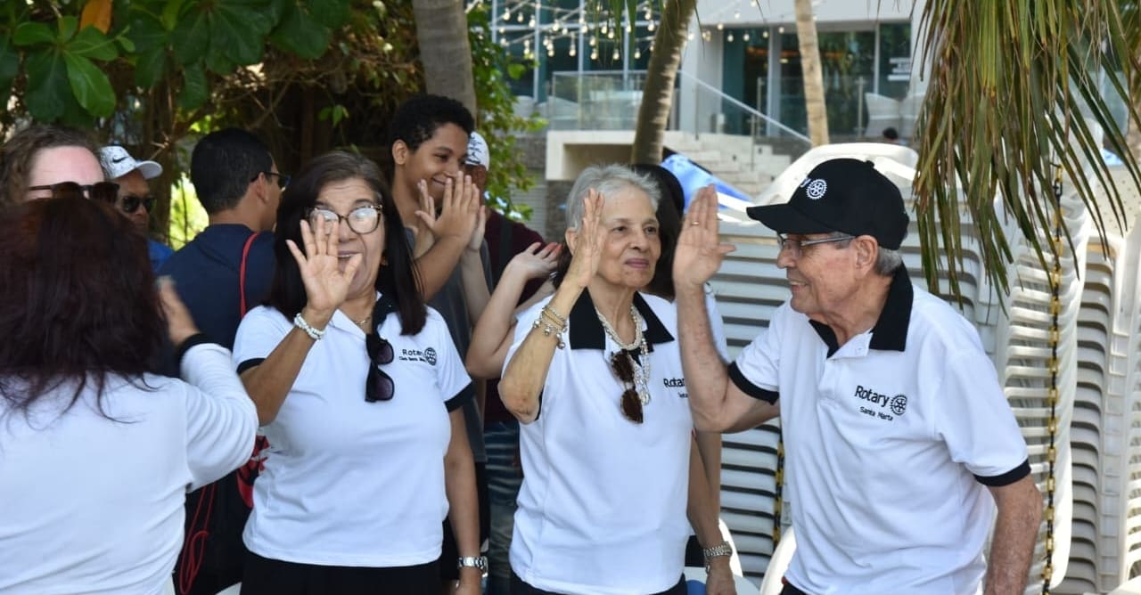 A group of people in matching white shirts and Rotary hats and logos, representing the Bellevue Breakfast Rotary Club, stand outdoors, smiling and giving high fives, with trees and stacked chairs in the background.
