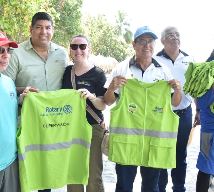 A group of people smile outdoors, with trees in the background. Two men in front hold bright green vests labeled Rotary Club de Soto la Marina Supervisor, proudly supporting our causes. Others stand nearby, some wearing Rotary shirts and hats.
