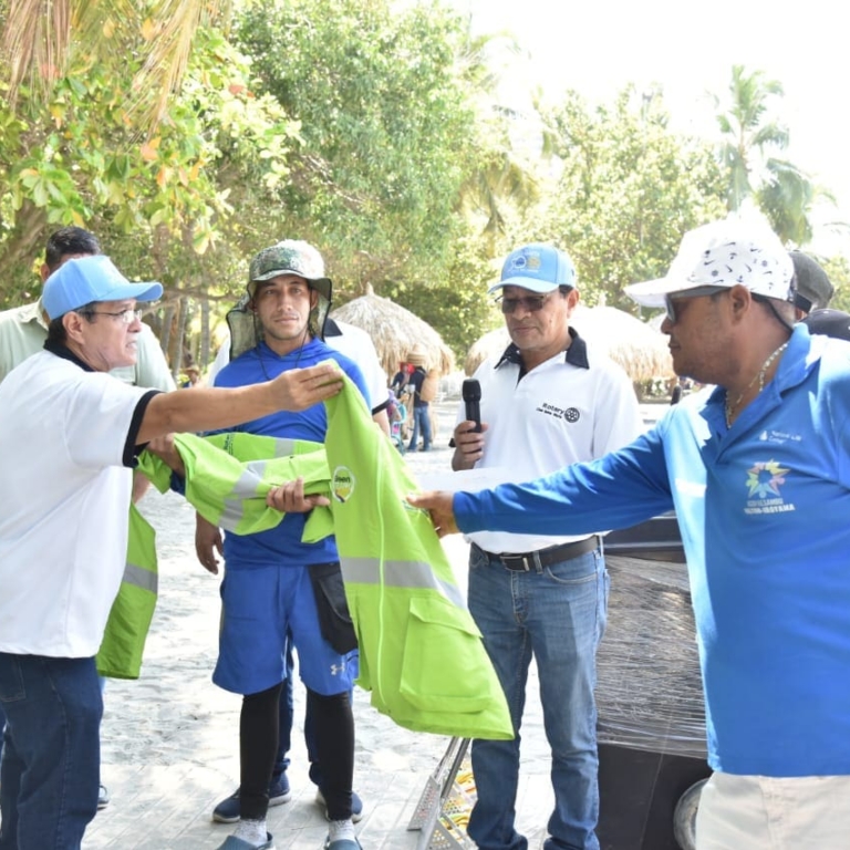 Five men stand outdoors in the shade, handing out bright green safety vests in support of our causes. One man holds a microphone, while the others participate in the distribution. Trees and sunlight are visible in the background.