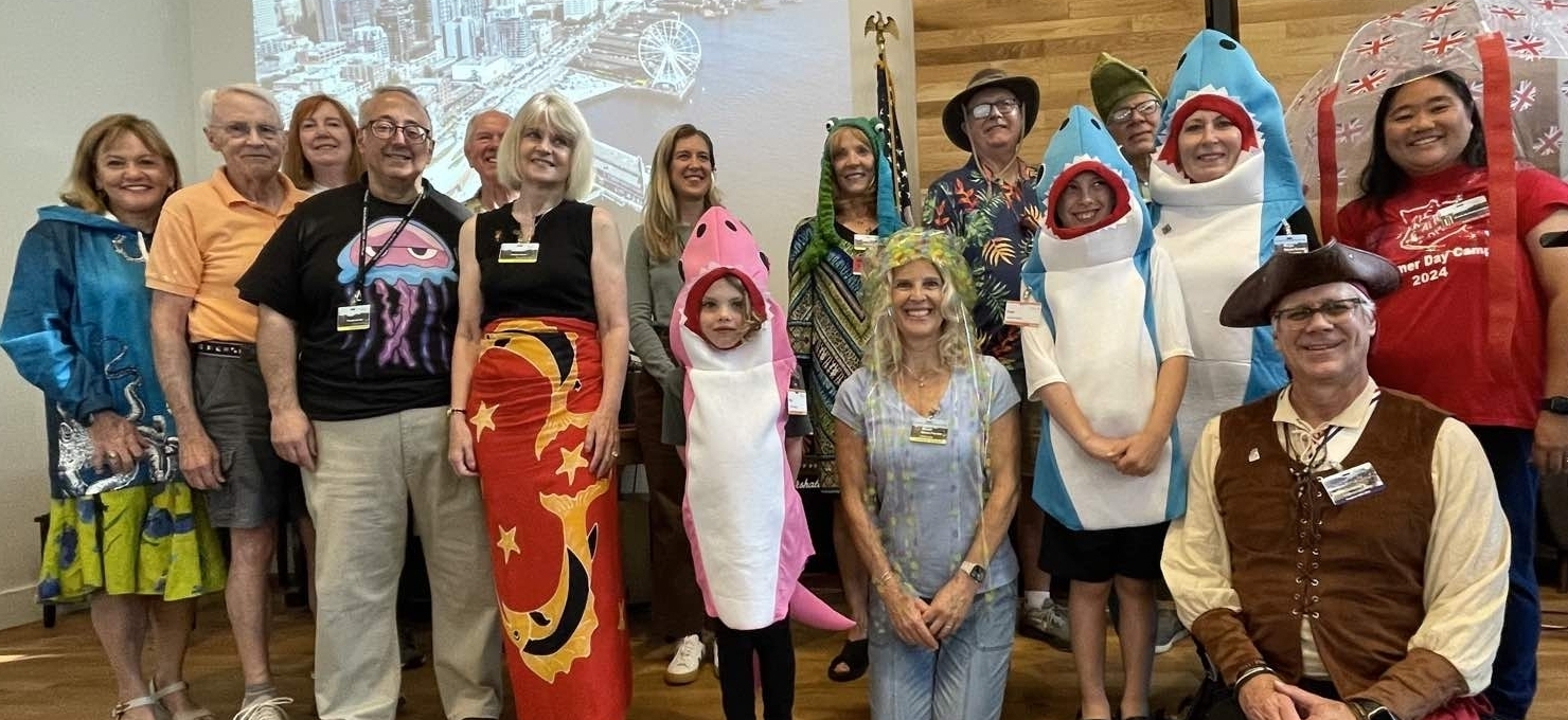 A diverse group of people stand in a line, many wearing costumes—including shark and frog hoodies, a pirate outfit, and a Union Jack umbrella hat—posed for a group photo indoors with a projection of a cityscape in the background, united in celebration of our causes.