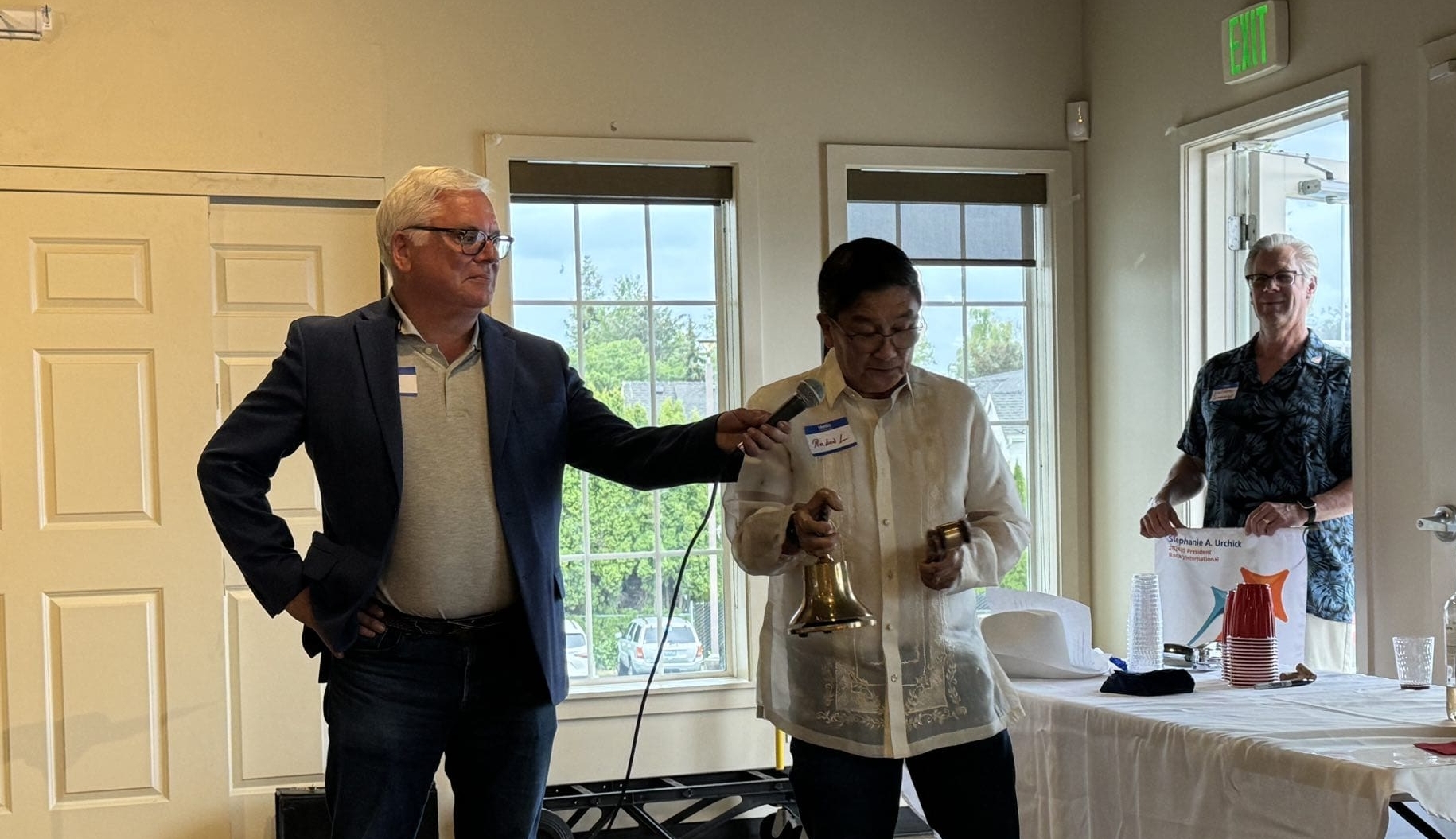 Two people stand indoors; one holds a microphone for the other, who is ringing a bell and holding a gavel—perhaps signaling the start of an event focused on international service projects. In the background, a third person stands by a table with drinks and snacks, preparing for the gathering.