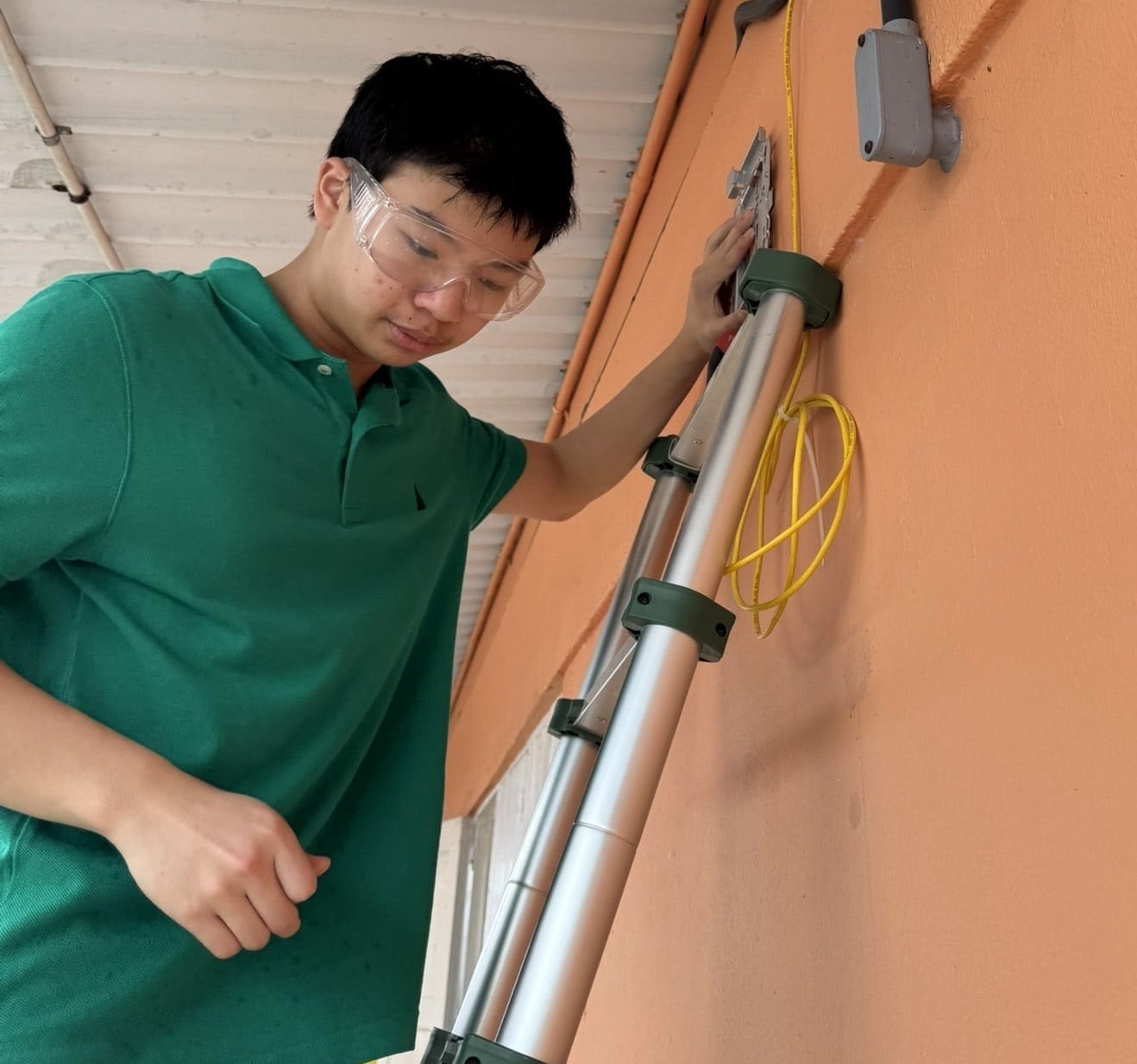 A person in a green shirt and safety goggles stands on a ladder, working on an orange wall with yellow cables and an electrical box—demonstrating their dedication to our causes through hands-on work and attention to safety.