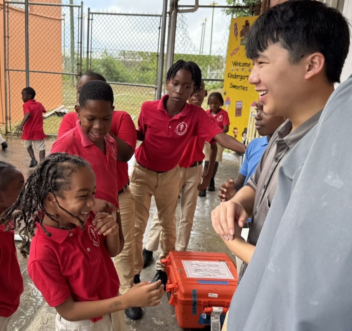 A group of children in red shirts and khaki pants laugh and interact with a smiling young man outside near a fence, enjoying a lively moment together during an event supported by the Bellevue Breakfast Rotary Club.