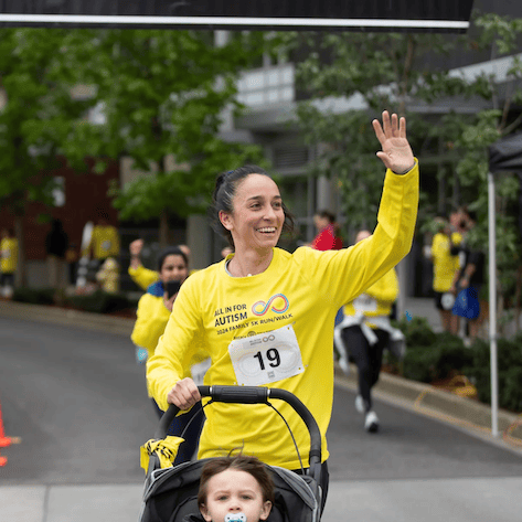 A woman in a yellow All In For Autism shirt smiles and waves while pushing a child in a stroller during a race supporting local service projects. The child has a pacifier. Other runners in yellow shirts are visible in the background.