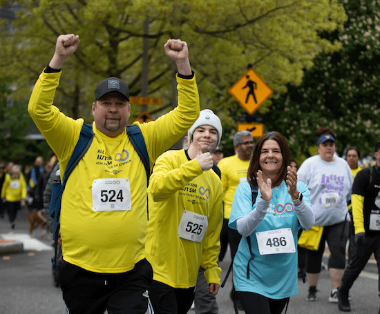 Three people at a walk event, smiling and celebrating. Two men in yellow shirts, one with arms raised, and a woman in a blue shirt are in the foreground with other participants in the background, coming together to support club donations and community spirit.