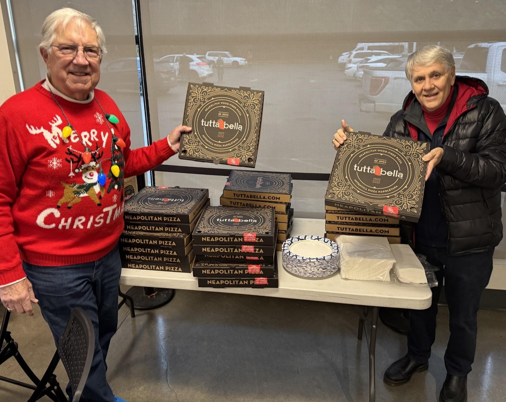 Two older men stand by a table with several pizza boxes and paper plates. One, who wears a festive Christmas sweater and smiles warmly, chats with his friend in the dark jacket as he holds a pizza box—making it the perfect moment to visit the club for some holiday cheer.