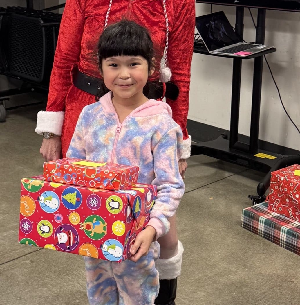 A young child in a colorful onesie holds two wrapped presents and smiles, her joy reflecting the spirit of club donations during the holidays. An adult in a red festive outfit stands behind her, with holiday gift boxes and a TV in the background.