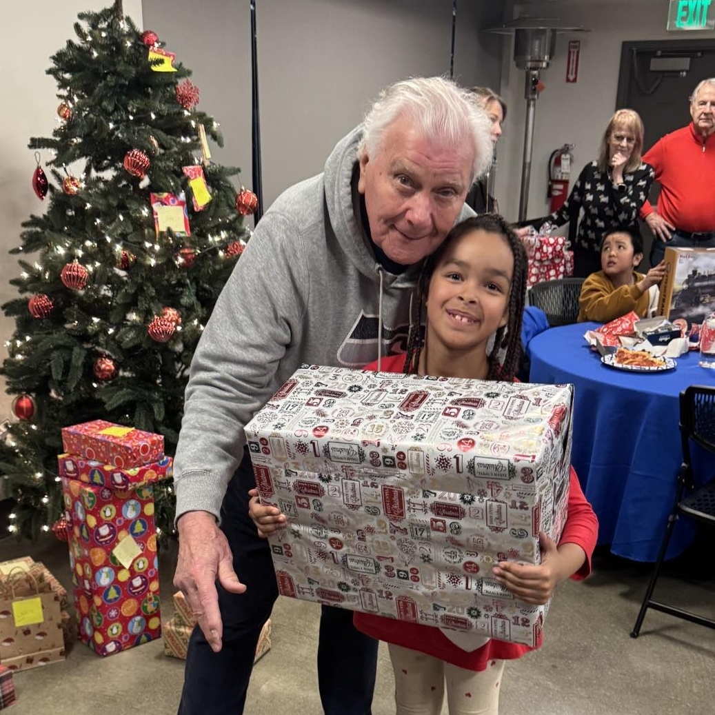 An older man hugs a smiling child holding a large gift box. They stand near a decorated Christmas tree with wrapped presents, celebrating the joy that comes from supporting our causes. People sit at a table in the festive background, sharing in the warmth of giving and togetherness.
