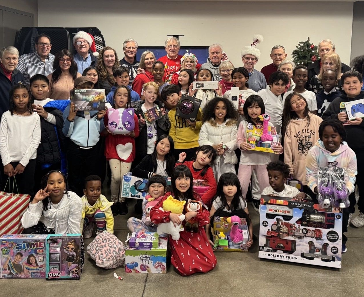 A large group of children and adults pose indoors, holding toys and gifts. A Christmas tree and holiday decorations are visible in the background. Everyone appears happy, celebrating a holiday gift-giving event that highlights our causes and the spirit of generosity within the community.