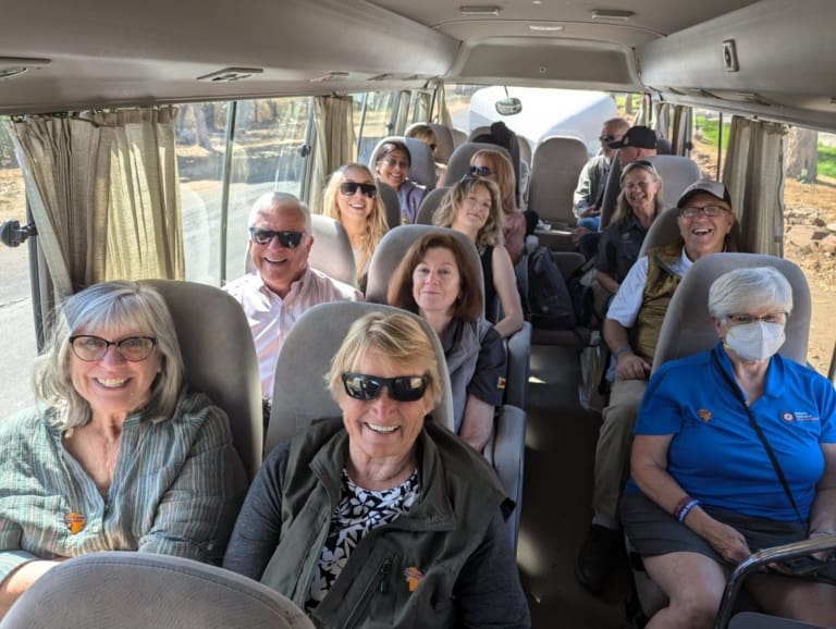 A group of smiling adults sits inside a small bus, enjoying a ride together while discussing their experiences with international service projects. One person wears a face mask, and sunlight streams through the windows. The mood is cheerful and relaxed.