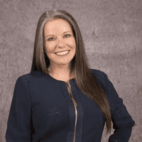 Woman with long straight brown hair wearing a dark blue zip-up top, smiling and standing in front of a textured gray background.