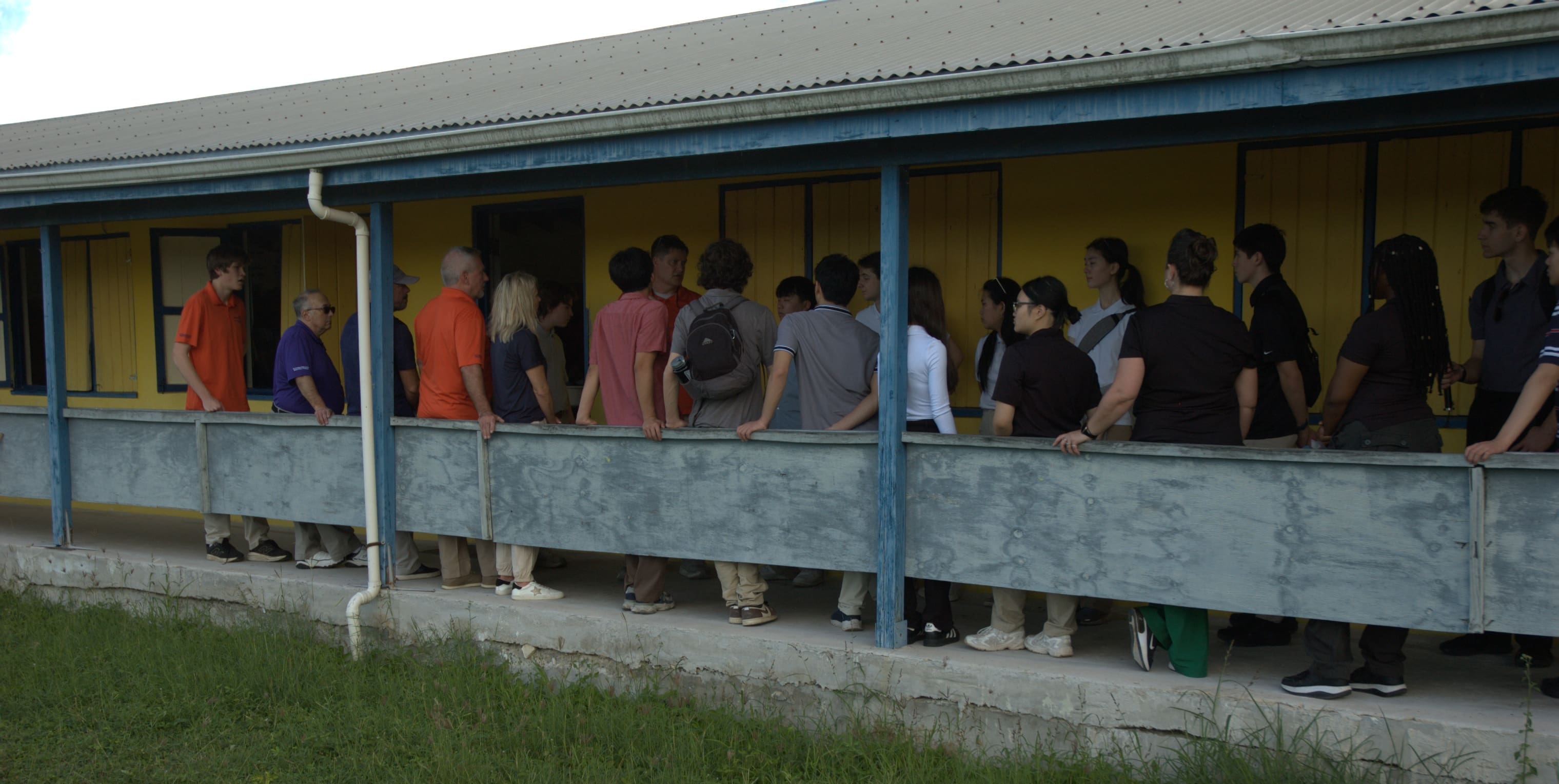 A group of people stand in line outside a yellow building with blue railings and a tin roof, waiting to enter through a door. Some are carrying backpacks, perhaps participating in an event organized by the Bellevue Breakfast Rotary Club.