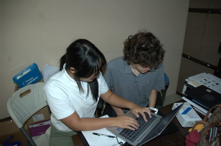 Two people sit at a desk, both focusing on a laptop. One is typing while the other looks at the screen, deep in discussion about international service projects. Papers, folders, and boxes are scattered around them, hinting at the scope and organization required for their global initiatives.