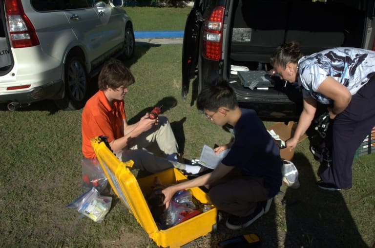 Three people work beside two parked SUVs; two sit on the grass sorting items from a yellow equipment case, while one stands at the vehicle’s open trunk organizing supplies for international service projects on a sunny day.