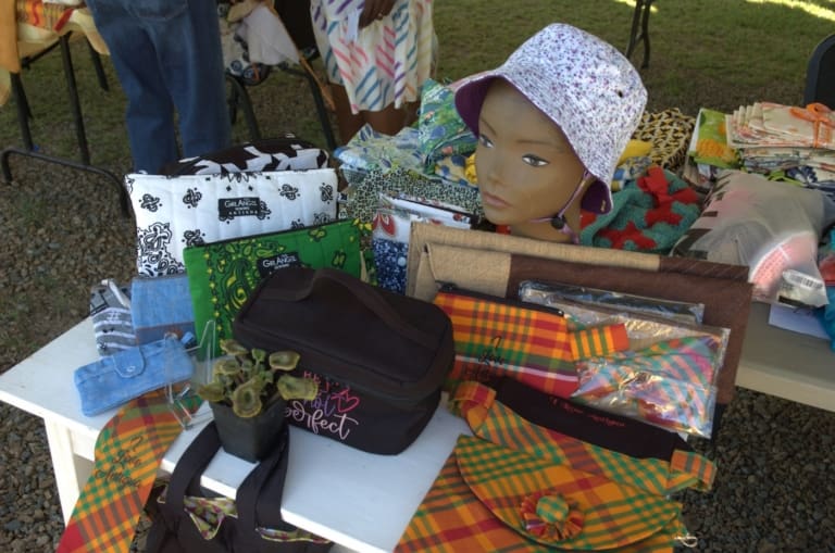 A display table with colorful bags, pouches, a mannequin head wearing a floral hat, a small potted plant, and various patterned fabric items at an outdoor market showcases the vibrant handiwork often featured in international service projects.