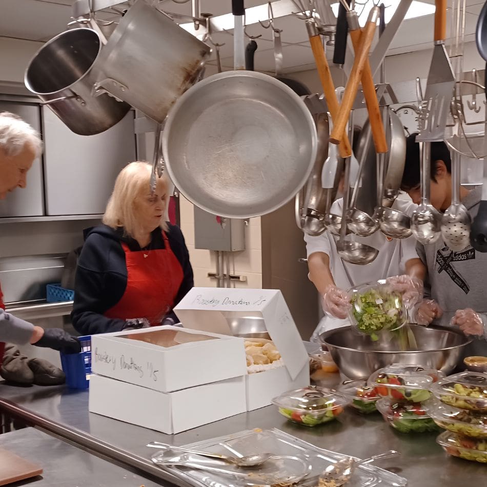 Four people work together in a kitchen, preparing salads and boxed food as part of local service projects. Kitchen utensils hang above, and various salads and meal boxes are arranged on the stainless steel counter.