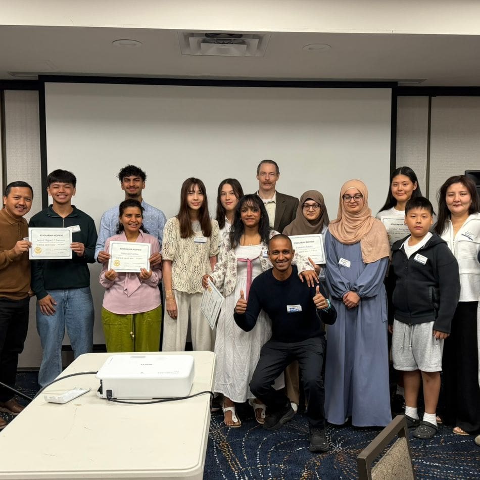 A group of 14 people of diverse ages and backgrounds pose indoors. Several hold certificates and smile at the camera, standing in front of a large projection screen in a conference room, celebrating their involvement in local service projects.