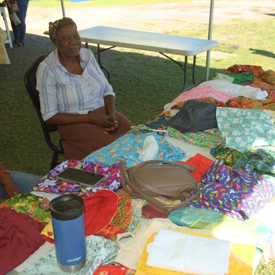 An older woman sits at a table displaying colorful handmade clothes and fabric items outdoors, showcasing the creativity often found in international service projects. A tumbler, phone, and bag are on the table. The scene is under a tent with grass visible in the background.