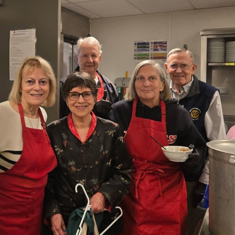 Five older adults smile in a kitchen. Three women in red aprons stand in front, one holding a bowl, another holding hangers. Two men stand behind them near a large pot. The setting appears to be a Bellevue Breakfast Rotary Club community or volunteer event.