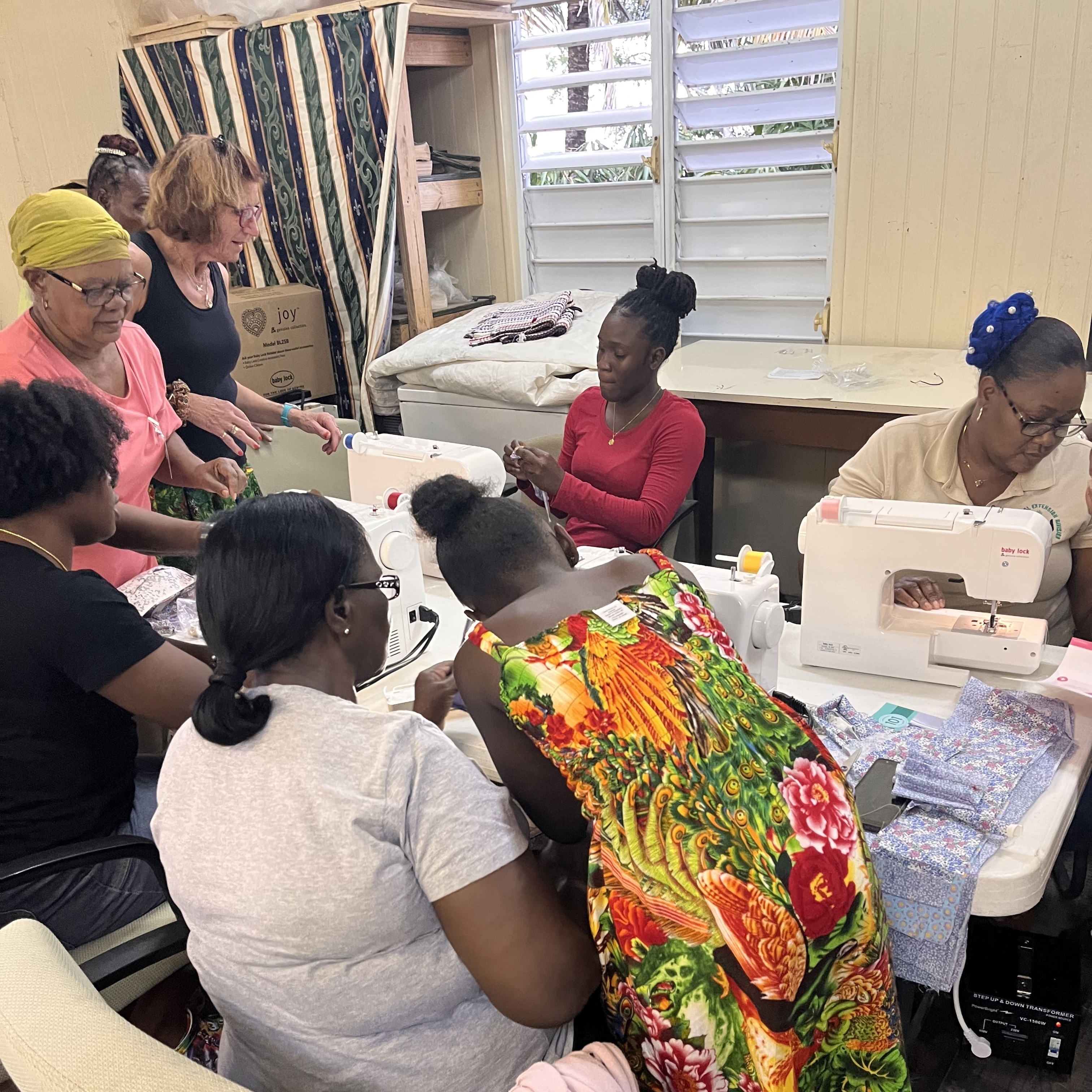 A group of women, some seated at sewing machines and others standing, work together in a room with colorful fabrics and sewing supplies on the tables, united by their dedication to our causes.