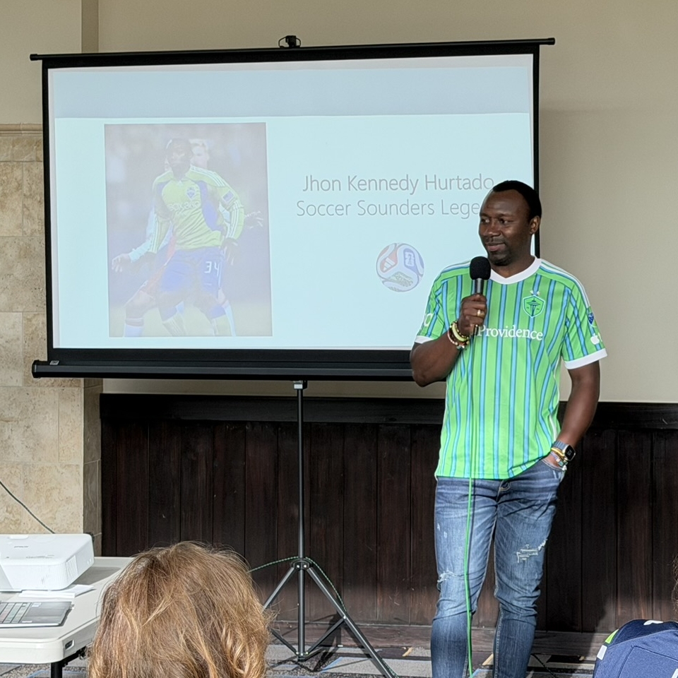 A man wearing a green soccer jersey stands with a microphone beside a screen displaying “Jhon Kennedy Hurtado Soccer Sounders Legend” and his photo, inviting others to join the club. Audience members are seated, watching the presentation.