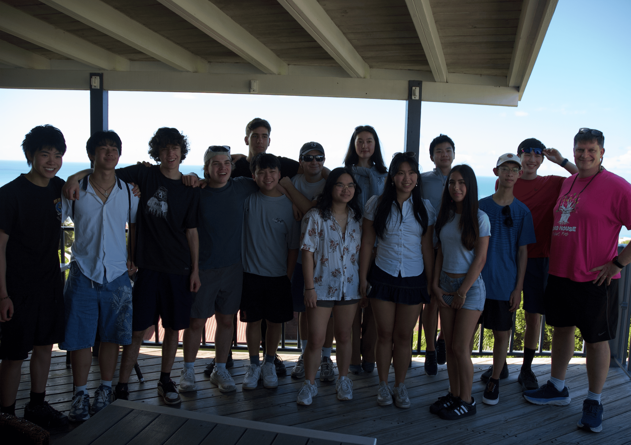 A group of 16 people stands together on a wooden deck under a roof, smiling at the camera with an ocean view and blue sky in the background—inviting you to join the club and be part of their vibrant community.