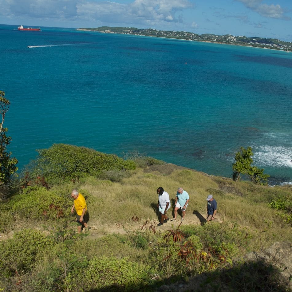Four people hike up a grassy hill overlooking bright blue ocean water, with a distant shoreline and a red ship visible under a partly cloudy sky—an adventure that invites you to join the club and experience the beauty together.