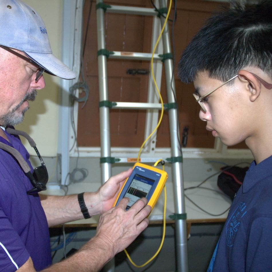 An adult shows a teenager how to use a handheld electronic device, with a yellow cable and a ladder in the background—evoking the hands-on learning often seen during international service projects.