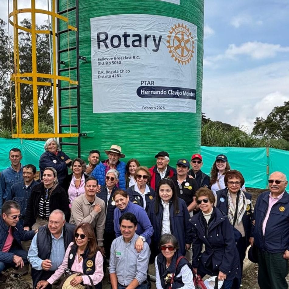 A large group of people pose in front of a green water tank with a large Rotary Club sign, highlighting their commitment to international service projects. Some are standing, others are seated, and they are all smiling outdoors on a cloudy day.