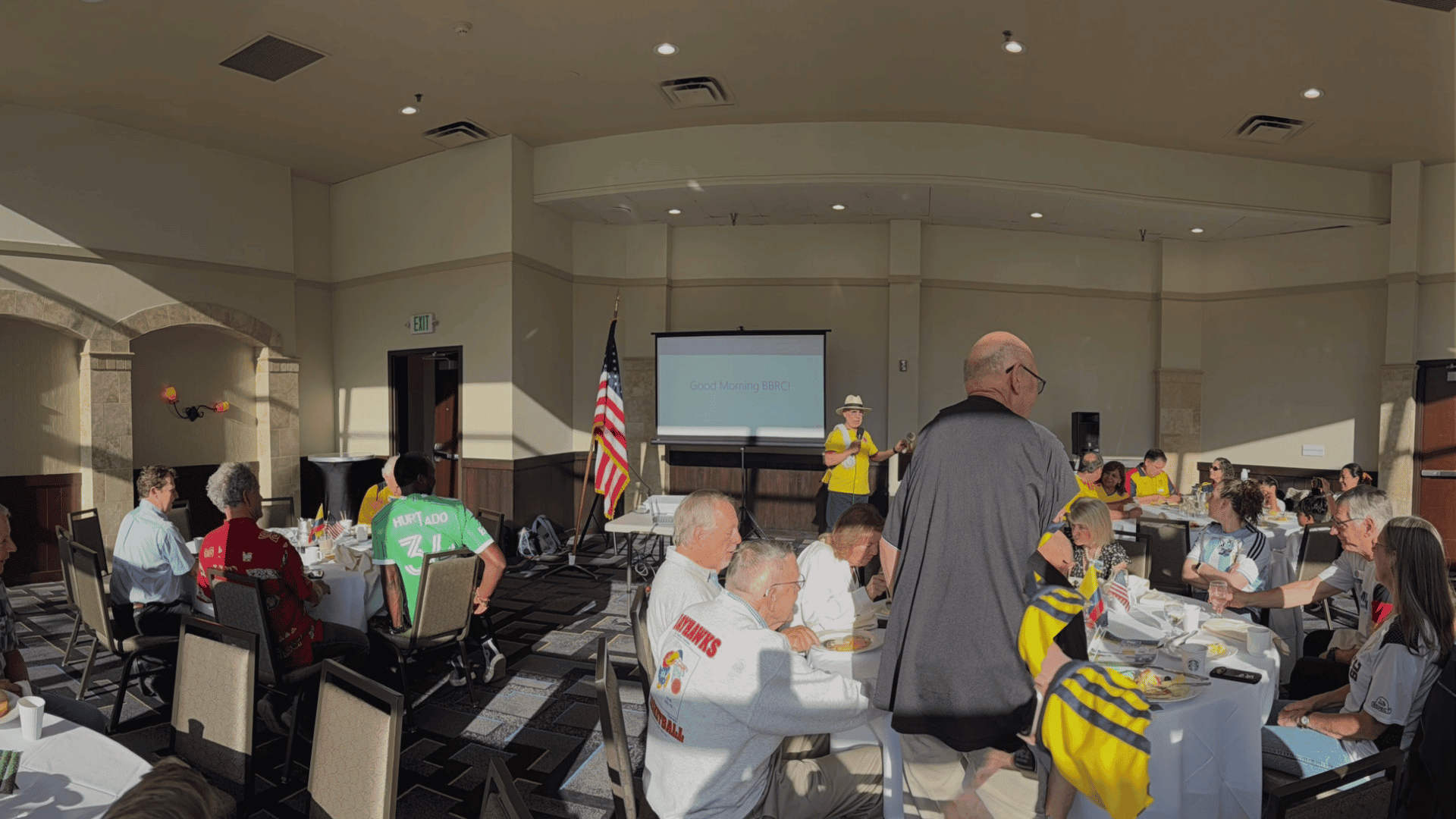 People sit at round tables in a bright meeting room with food, facing a screen displaying Guest Rotarian (BRC). An American flag stands near the screen; a man in a yellow vest speaks at the front, inviting attendees to visit the club and learn more about its activities.