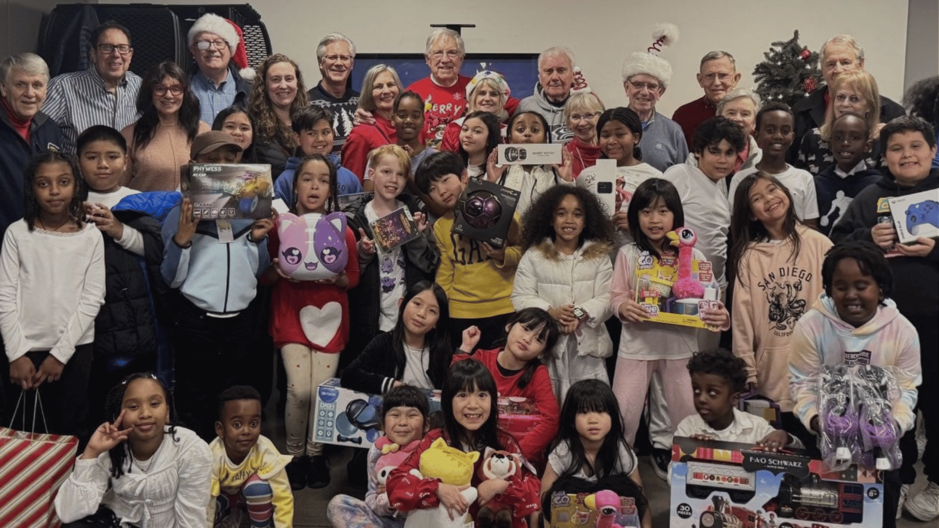 A large group of adults and children pose together indoors, some wearing festive holiday attire. Many children hold toys and gifts from recent local service projects, and everyone is smiling, creating a joyful, celebratory atmosphere.